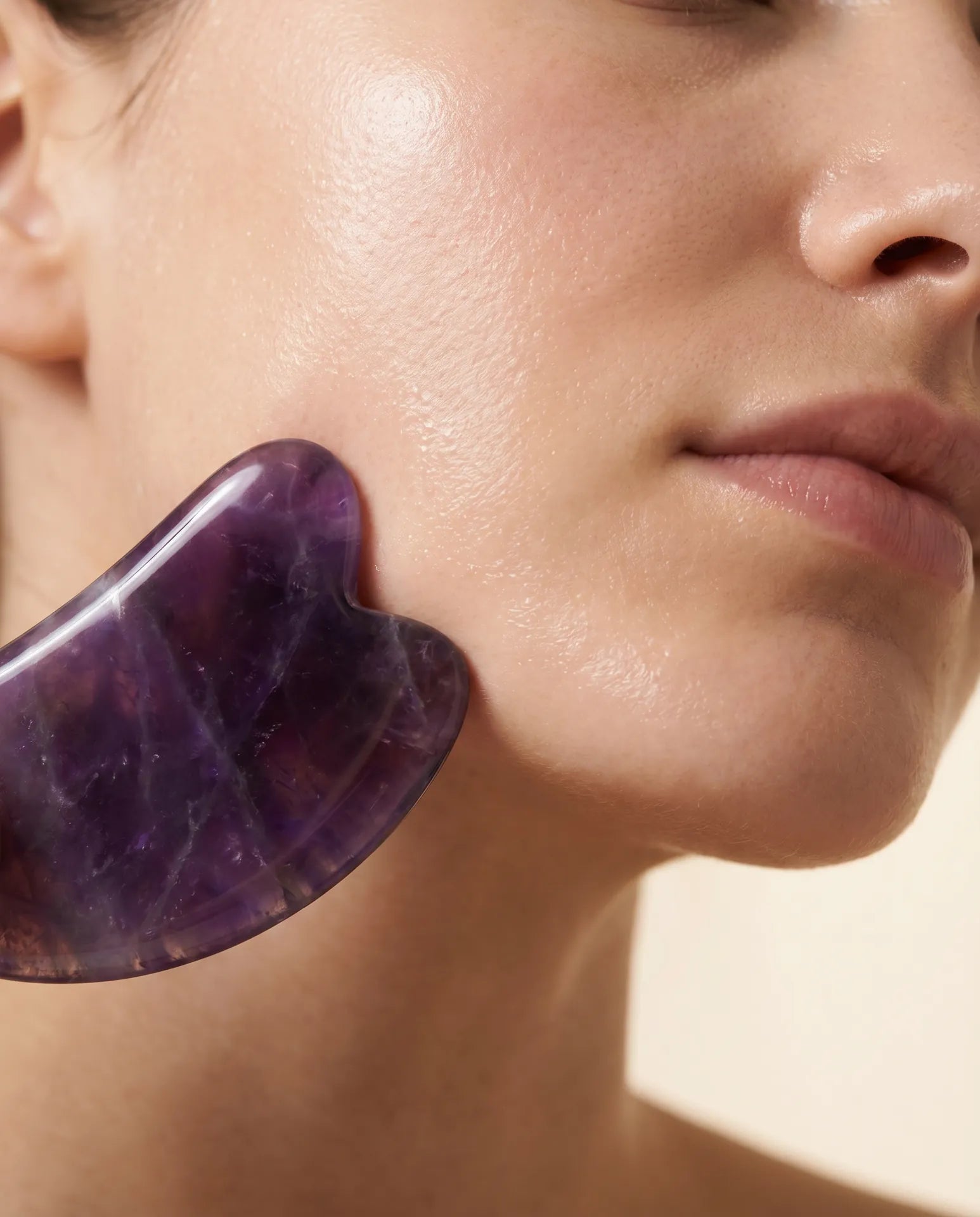 A polished amethyst gua sha stone gliding along the curve of a cheek and jawline in soft natural light