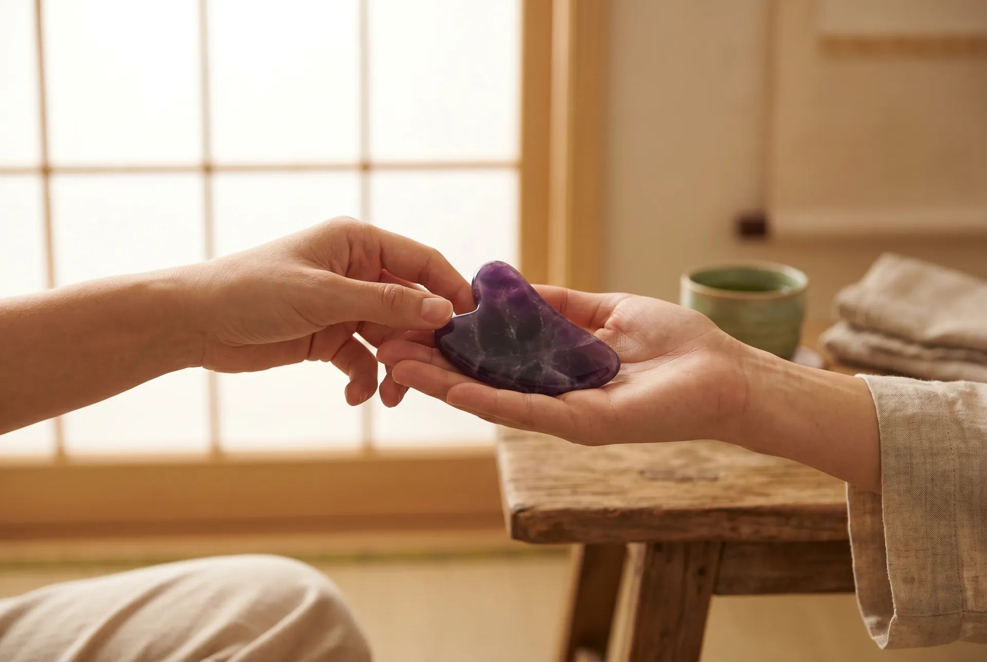 Two hands meeting in soft afternoon light, one offering a polished amethyst gua sha stone, the other reaching to receive it